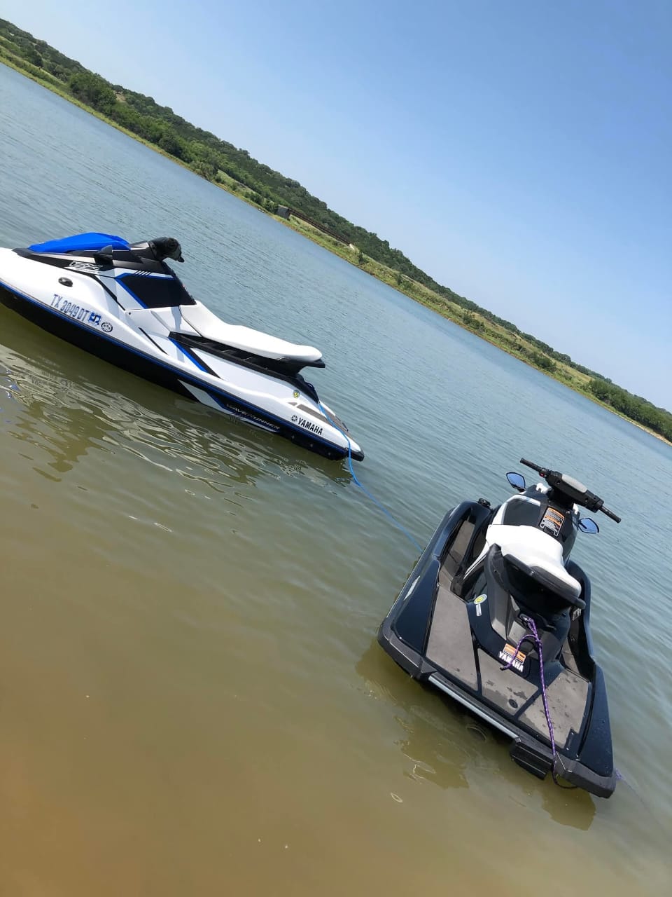 Two jet skis floating in the water near a grassy and wooded area below open sky. The left jet ski has a white and blue hul...
